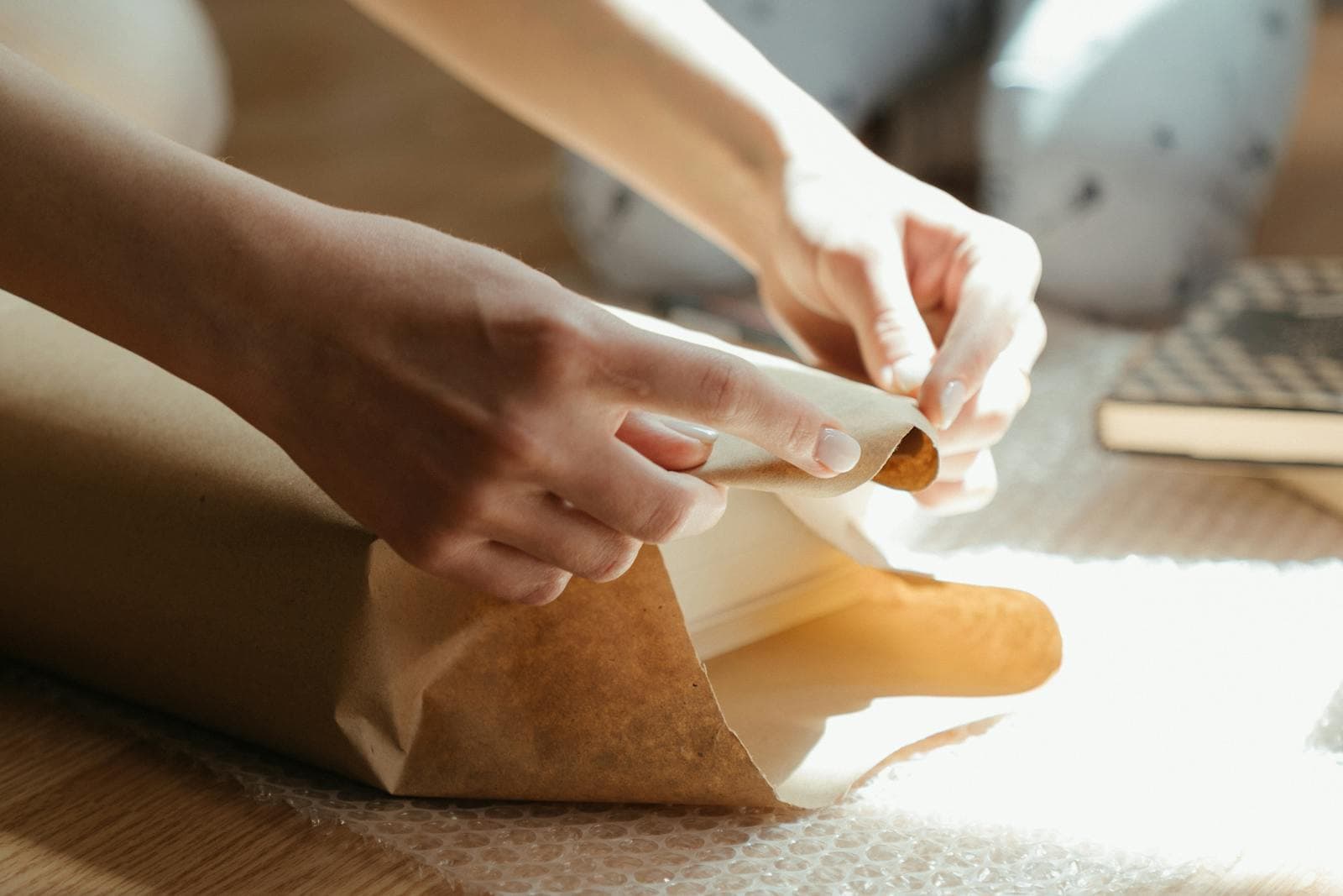 Two hands carefully wrap fresh food in brown kraft paper in quiet, warm light.